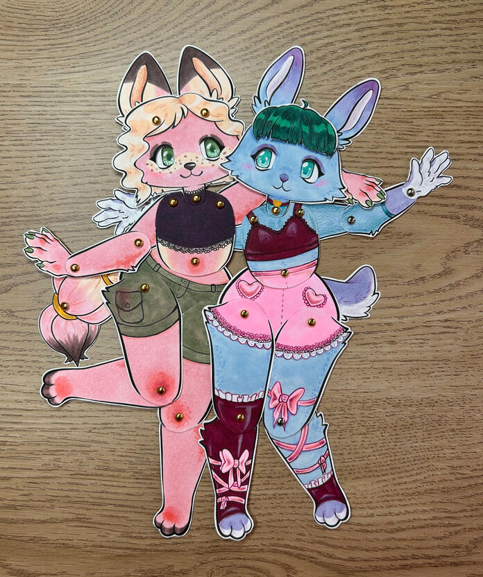 Two paper dolls, made using the kit. they are posed with their arms around each other.